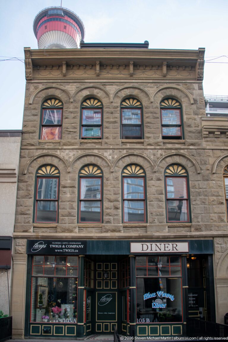 Three-story historic stone building with arched windows and diner storefront, with the Calgary Tower peeking above.