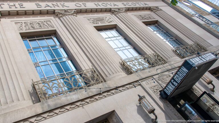 Upward view of the Bank of Nova Scotia stone façade with tall windows, pilasters, ornament, and metal railings.
