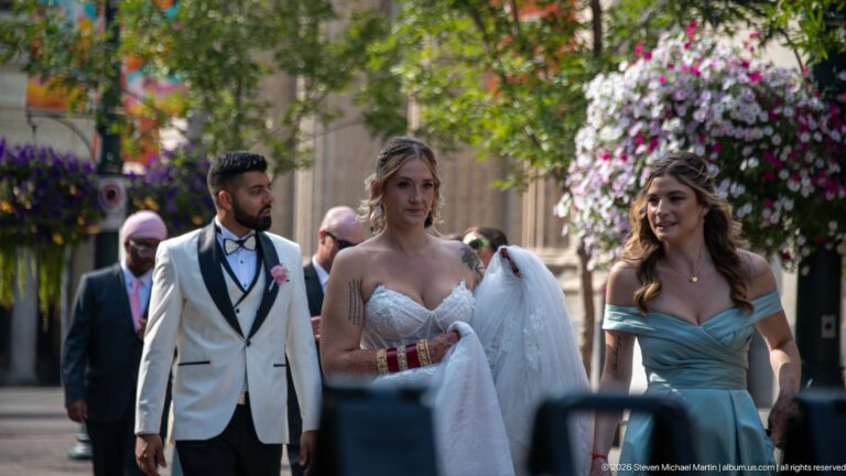 Bride, groom in a white jacket, and attendant in a blue dress walking along a flower-lined Calgary street.