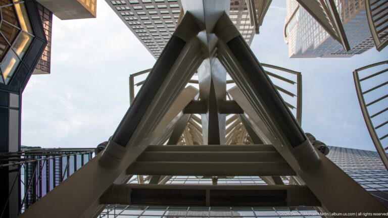 Upward detail of a white Galleria Tree’s tubular supports and slotted canopy framed by downtown Calgary towers.