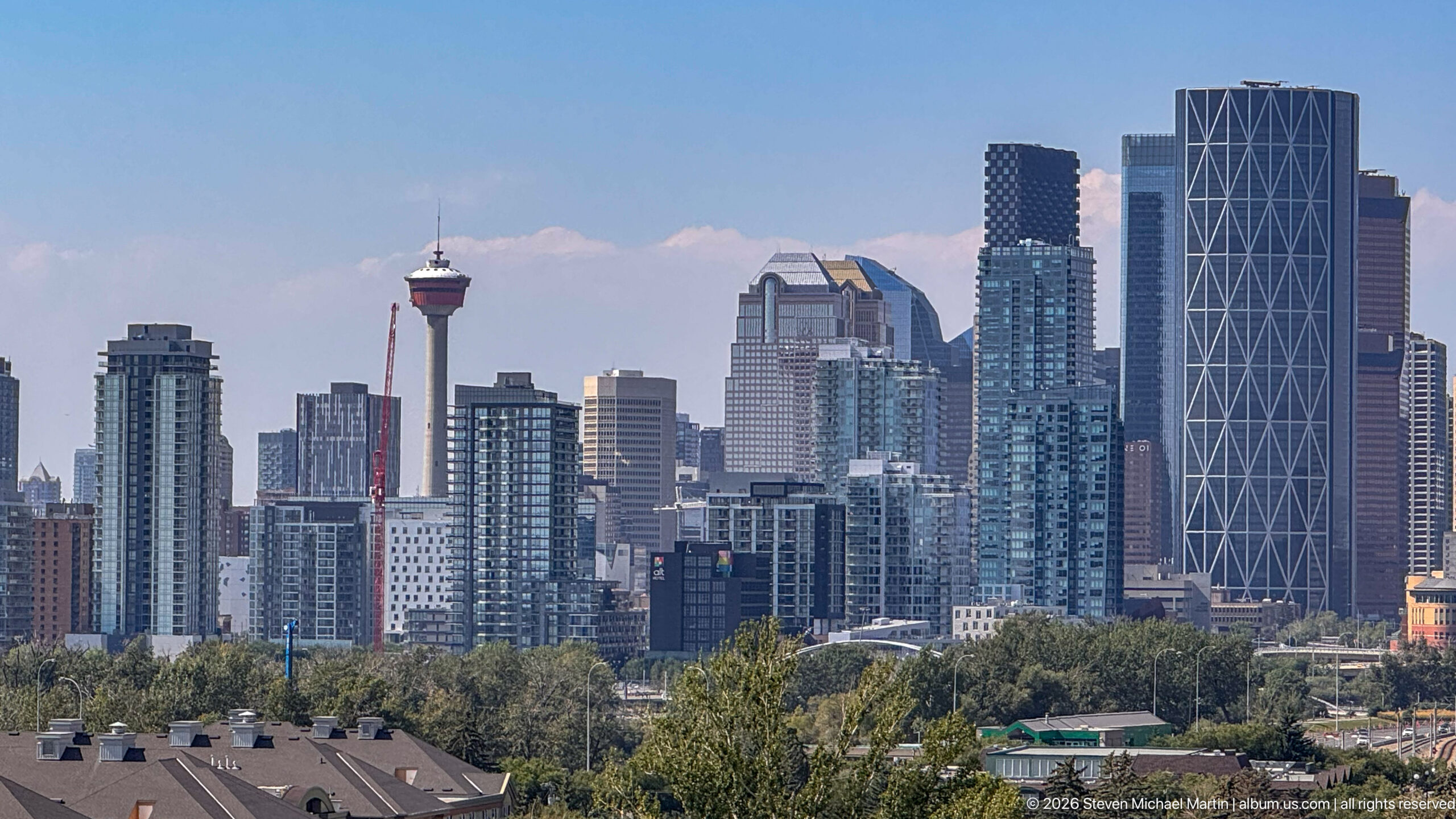 Calgary Tower amid dense downtown residential and office towers, viewed above trees and low buildings.