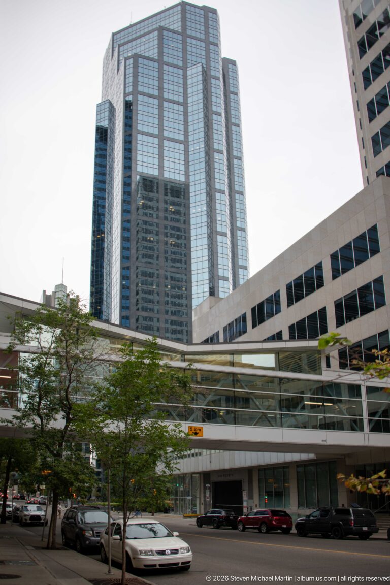 Downtown Calgary office towers and an enclosed elevated pedestrian walkway above a tree-lined street.