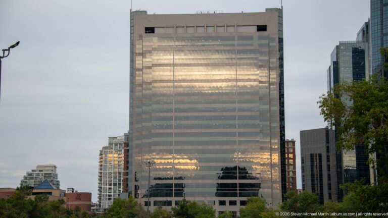 Large reflective glass office tower glowing with evening light above trees and smaller downtown buildings.