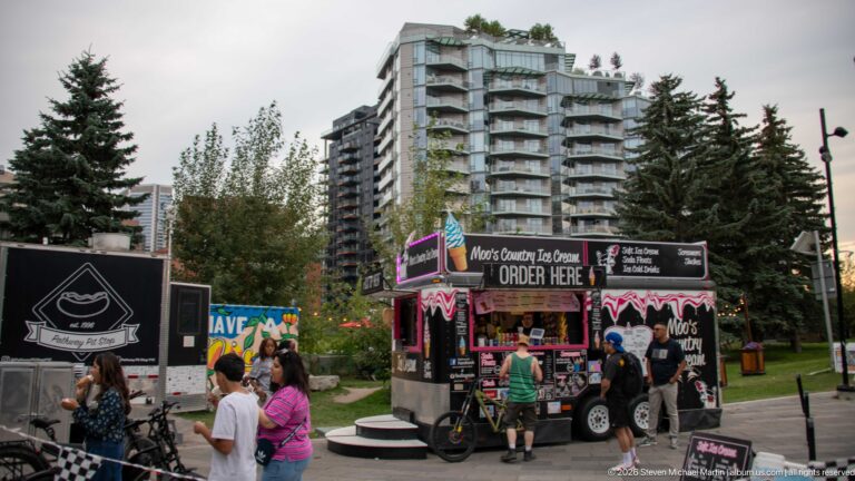 Customers gathered at colorful food trucks beside a Calgary park, with residential towers and trees behind.