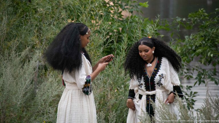 Two women in light dresses with dark trim standing among riverside vegetation, one looking at a phone.