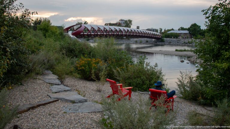 Red chairs beside a landscaped river path overlooking Calgary’s Peace Bridge across the Bow River.