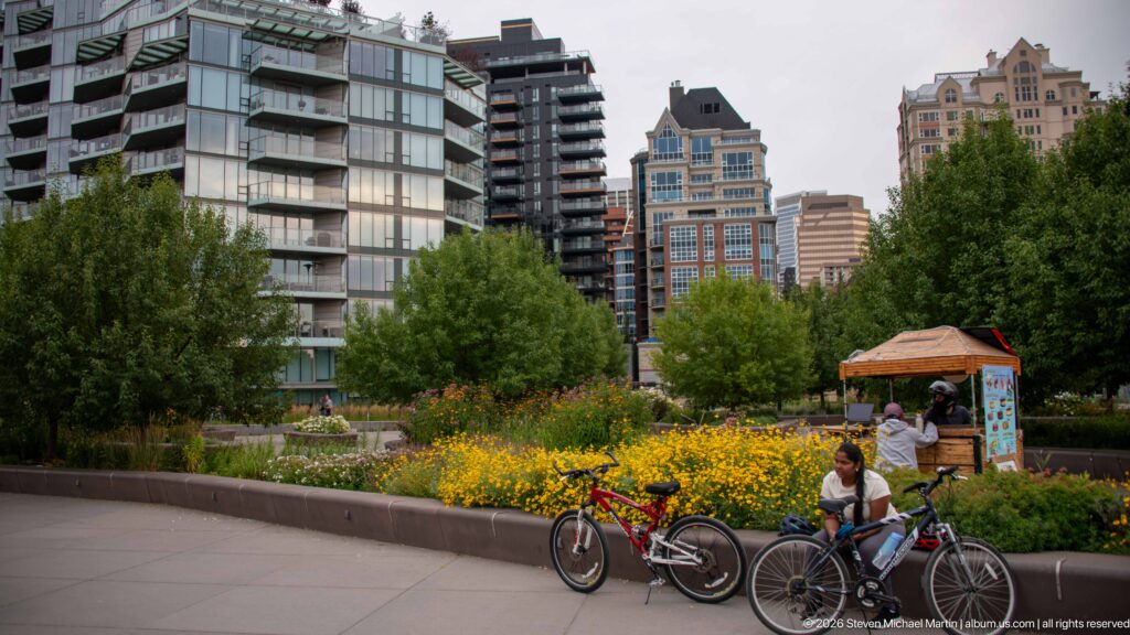 Cyclists beside yellow flower beds in a landscaped Calgary park with residential towers behind.