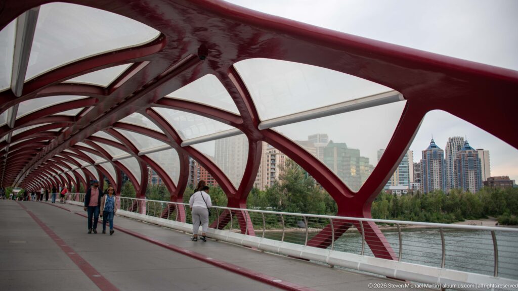 Red arches of Calgary’s Peace Bridge frame pedestrians, the river edge, trees, and downtown towers.