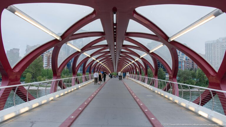 Centered view through Calgary’s Peace Bridge with repeating red arches, pathway lanes, lights, and pedestrians.