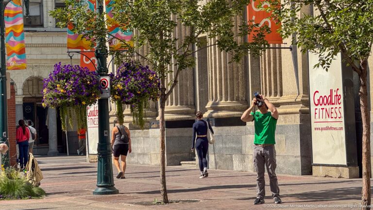 Man in green shirt photographing upward beside a fluted stone façade, hanging flowers, trees, and colorful street banners.