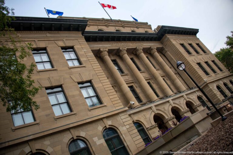 Low-angle corner view of McDougall Centre showing sandstone walls, paired columns, arched openings, attic windows, and Canadian flag.