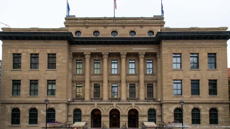 Frontal view of McDougall Centre with three arched entrances, paired columns, circular attic windows, sandstone façade, and rooftop flags.