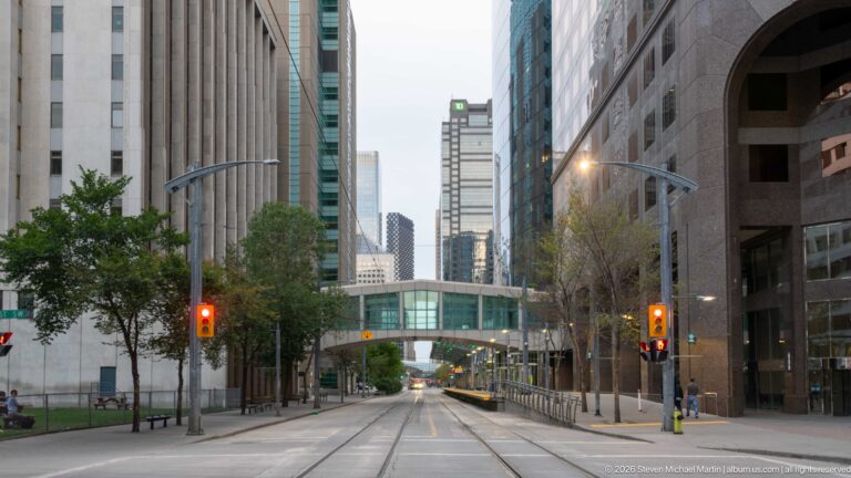 Quiet downtown Calgary street between office towers with red traffic lights and an enclosed pedestrian bridge ahead.