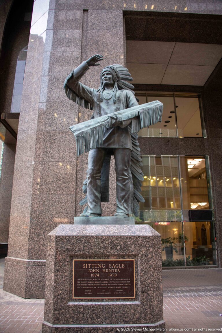 Bronze sculpture of Chief David Crowchild in feathered headdress and fringed clothing, shading his eyes, on a granite pedestal.