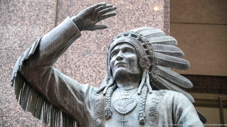 Close view of Chief David Crowchild bronze sculpture showing feathered headdress, raised hand, braids, necklaces, and detailed clothing.