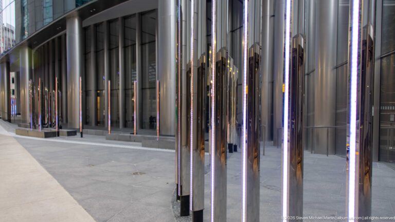 Rows of polished mirrored columns with vertical lights reflecting pedestrians and buildings along a modern Calgary sidewalk.