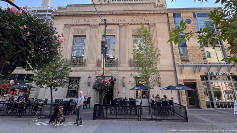 Historic Bank of Nova Scotia stone façade behind patio tables, umbrellas, hanging flowers, trees, and pedestrians.