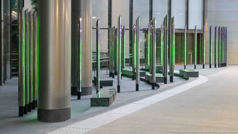 Rows of mirrored vertical columns glowing green beside large structural columns along a modern Calgary building.