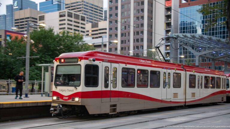 Red-and-white Calgary Transit CTrain traveling on downtown street-level tracks with high-rise buildings behind.