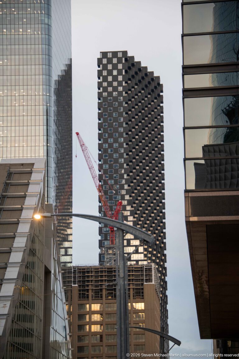 Dark balcony-covered high-rise under construction between downtown Calgary towers, with a red crane crossing the lower façade.