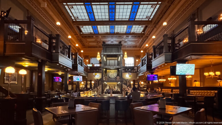 Wide historic bar interior with dark wood galleries, blue stained-glass ceiling panels, central clock, tables, and leather seating.