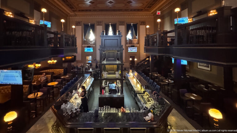 Elevated view down a long bar in an ornate two-level historic interior with dark galleries, lamps, and tall windows.
