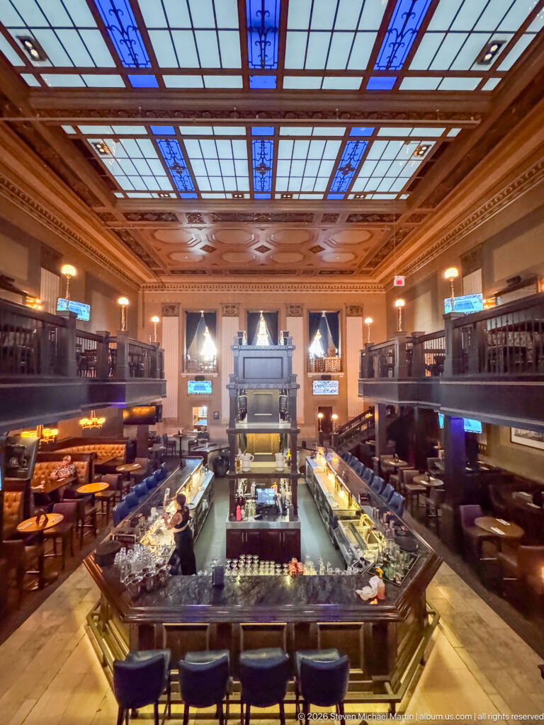 Wide elevated view of an ornate historic banking-hall bar with U-shaped counter, galleries, leather seating, and blue stained-glass ceiling.