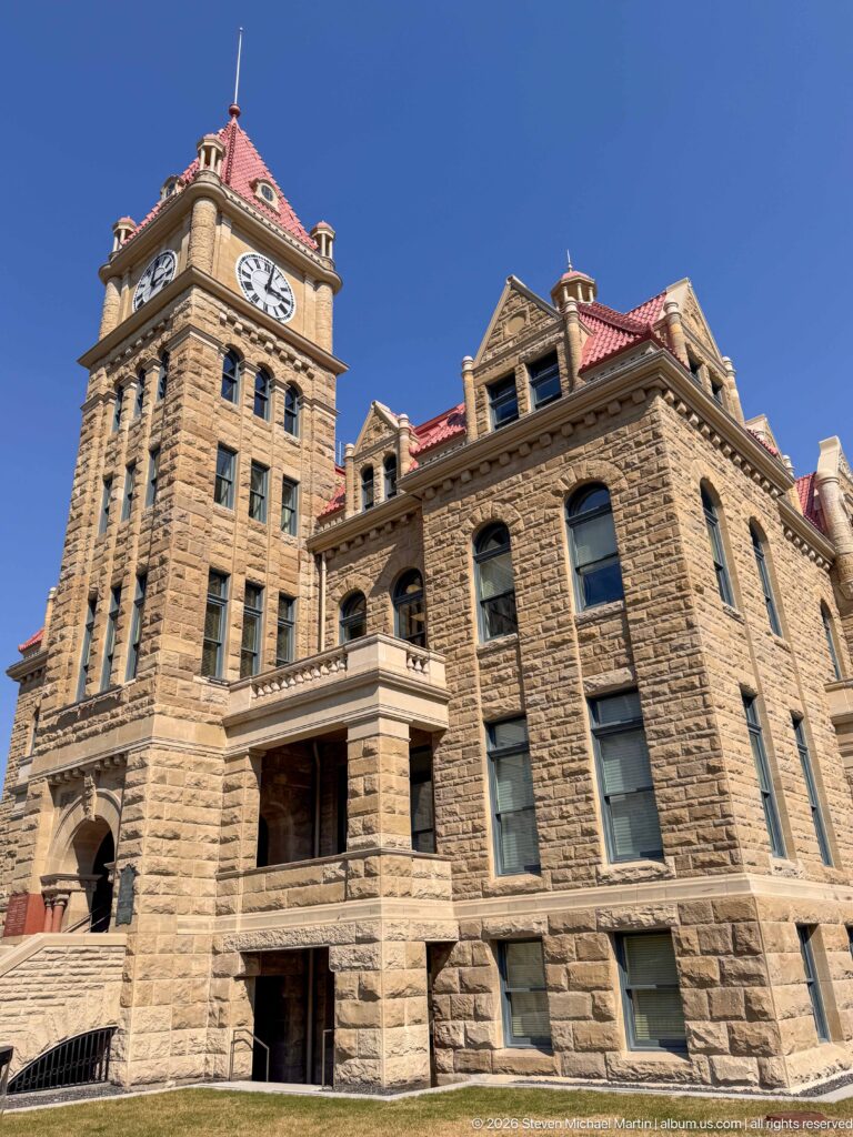 Historic Calgary City Hall with sandstone walls, clock tower, red roofs, arched windows, and projecting bays under blue sky.
