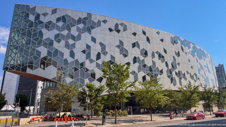 Wide street view of Calgary Central Library’s curved white geometric façade with trees, bicycle racks, and glazed opening.