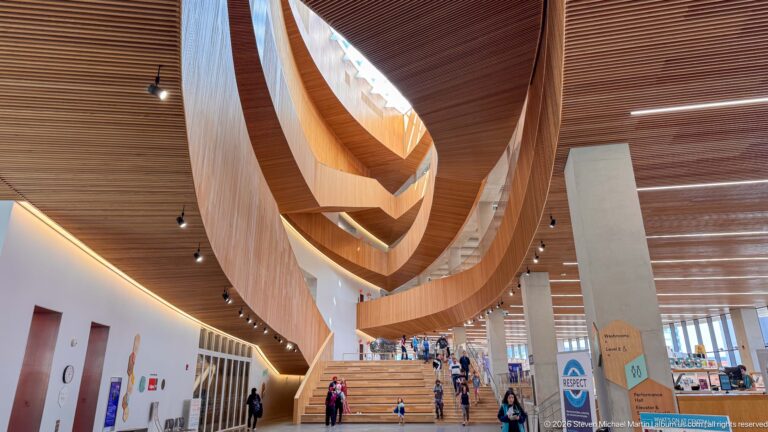 Calgary Central Library atrium with sweeping nested wood curves above a broad staircase and visitors.