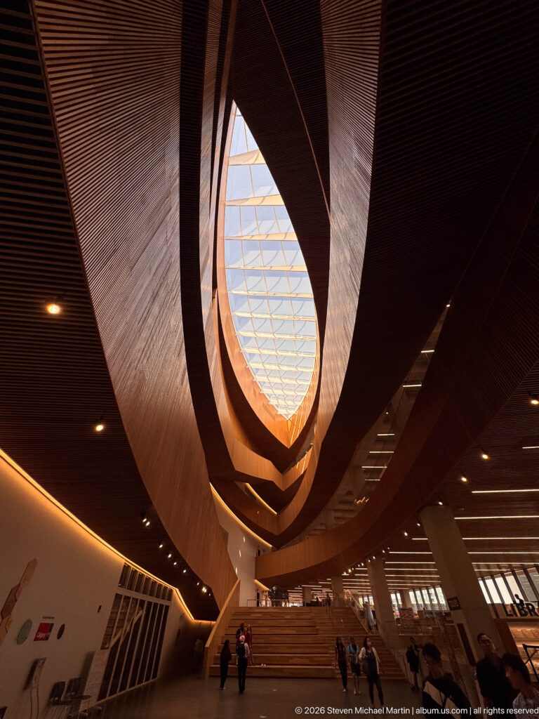 Low-angle view through Calgary Central Library’s layered timber atrium toward a skylight above the main stairs.