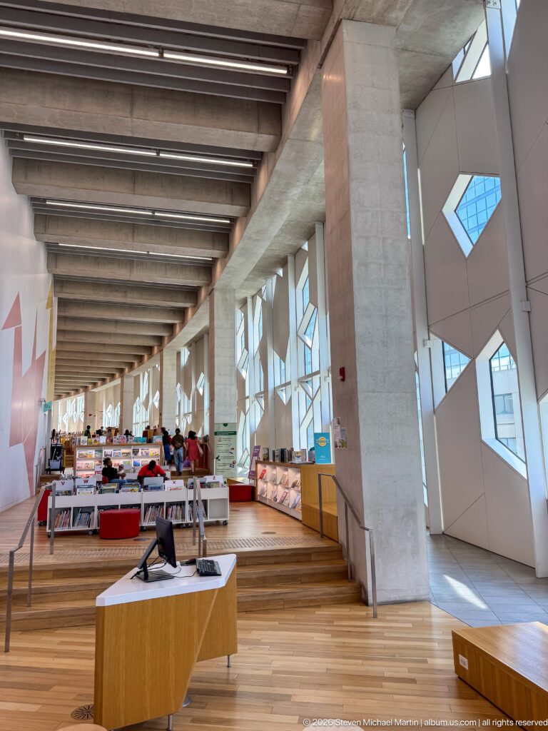 Open Calgary Central Library floor with low bookshelves, reading tables, colorful seating, concrete beams, and angular windows.
