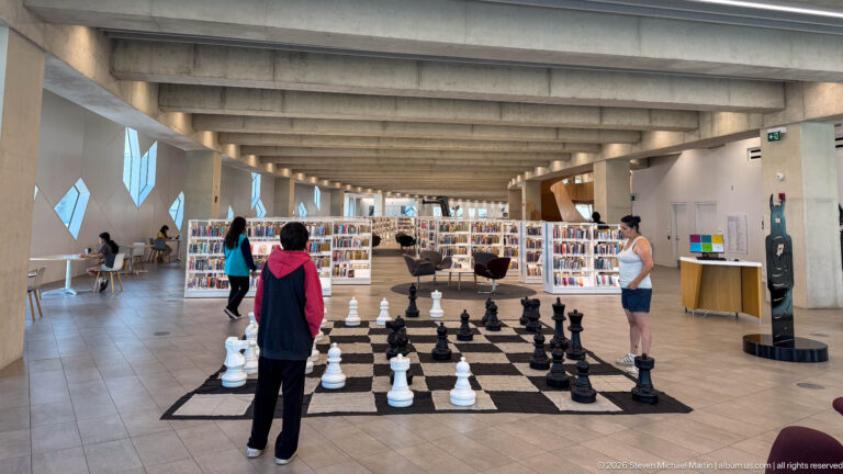 Visitors standing around a giant black-and-white chess set inside Calgary Central Library with bookshelves behind.