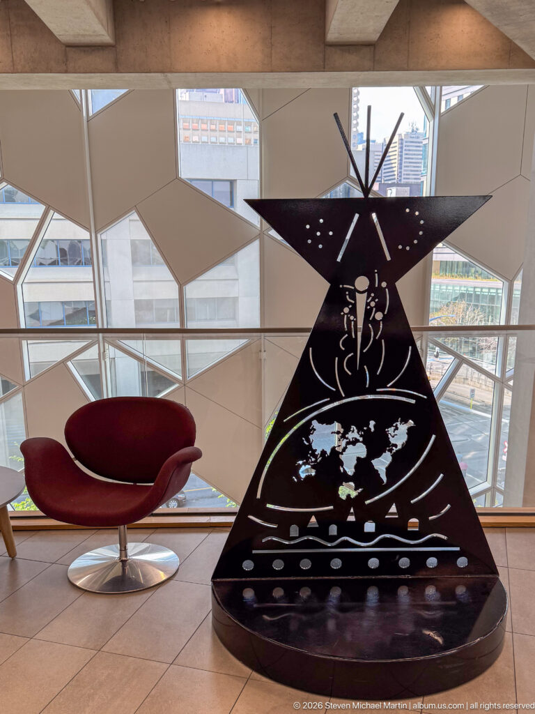 Black triangular sculpture with cutout figure, radiating lines, circles, and animal motifs beside a red chair.