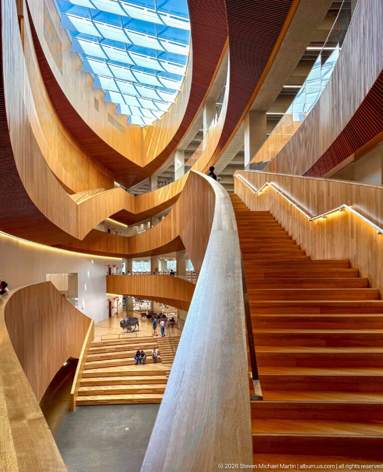 Wide staircase rising through curving wood-clad ramps and balconies inside Calgary Central Library beneath a skylight.