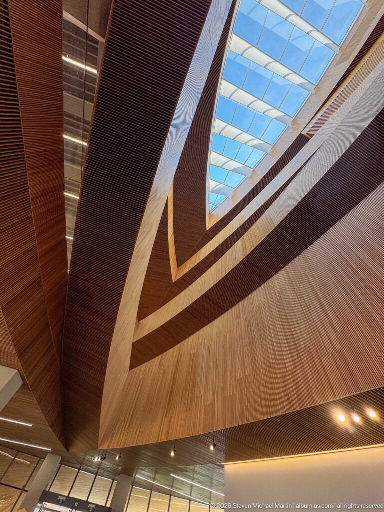 Upward view through Calgary Central Library’s curving timber balconies toward a bright skylight.