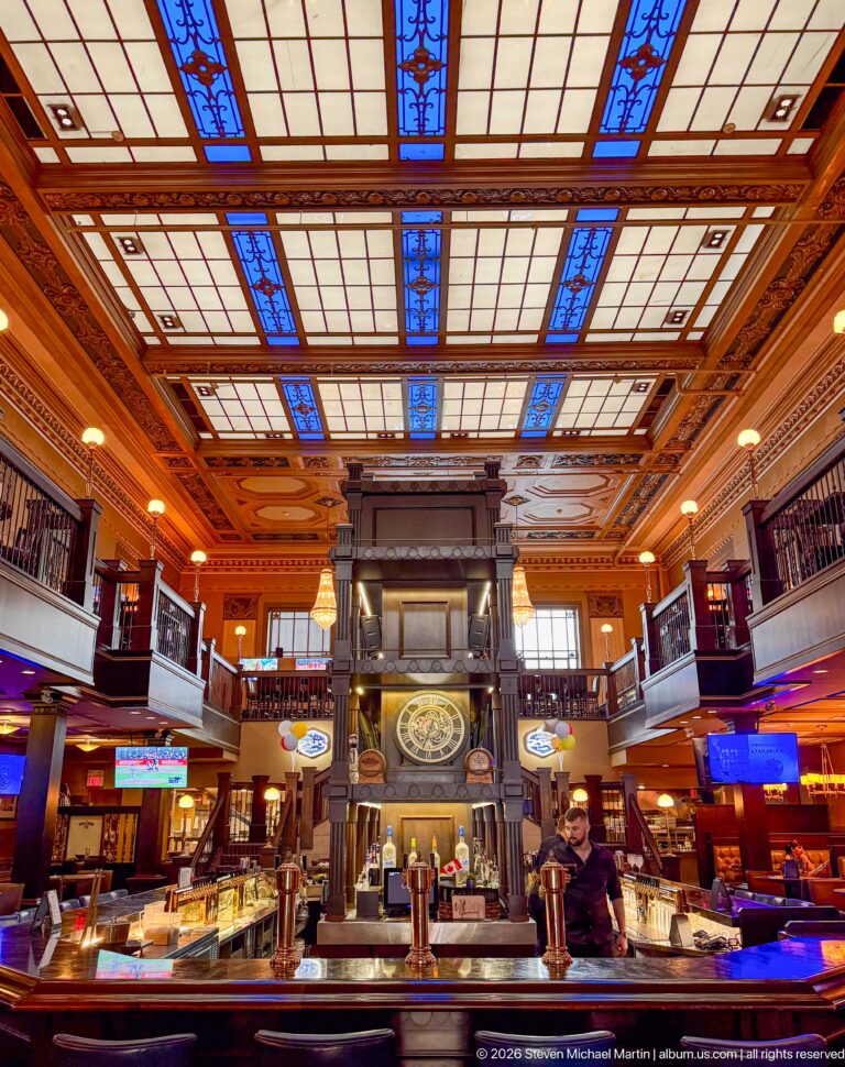 Symmetrical historic bar interior with central clock, long counters, dark galleries, and blue stained-glass ceiling panels.