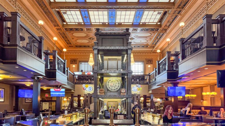 Ornate two-level bar with dark wood galleries, central clock, stained-glass ceiling panels, and warm lighting.