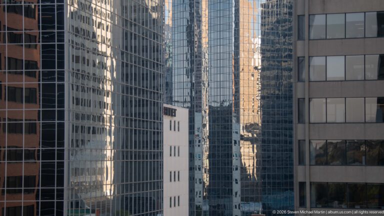 Tightly compressed view between reflective downtown Calgary office towers with mirrored façades and a narrow pale building centered.