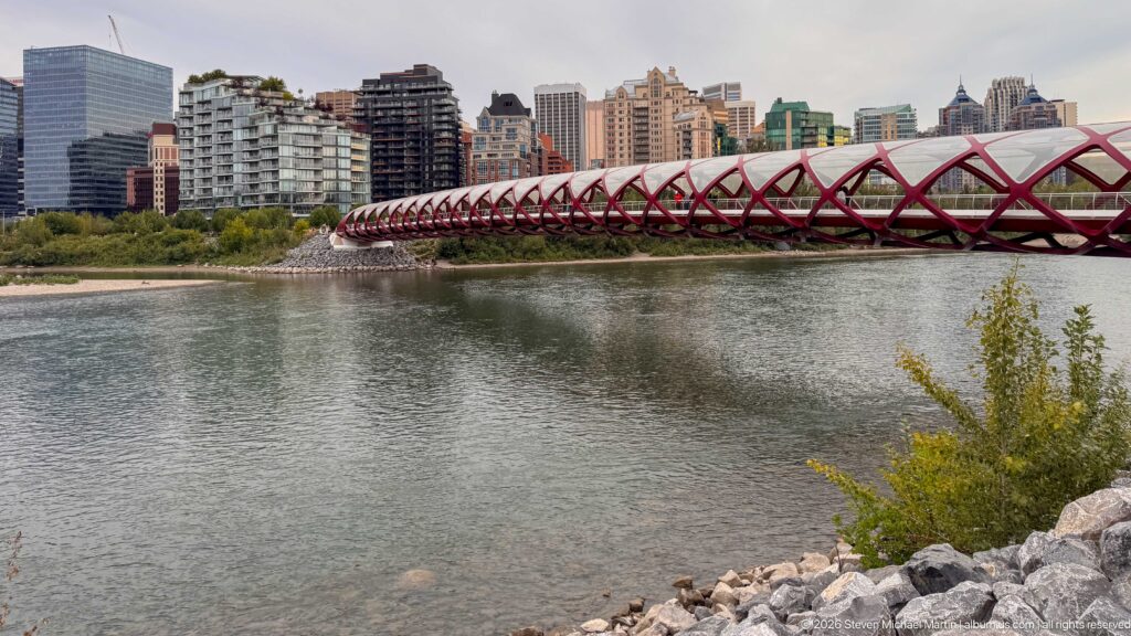 Calgary’s red Peace Bridge crossing the Bow River with residential high-rises behind and rocky shoreline in front.