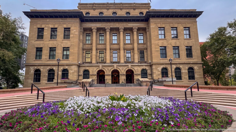Closer frontal view of McDougall Centre’s sandstone entrance bay, paired columns, three arches, broad steps, and colorful flower beds.