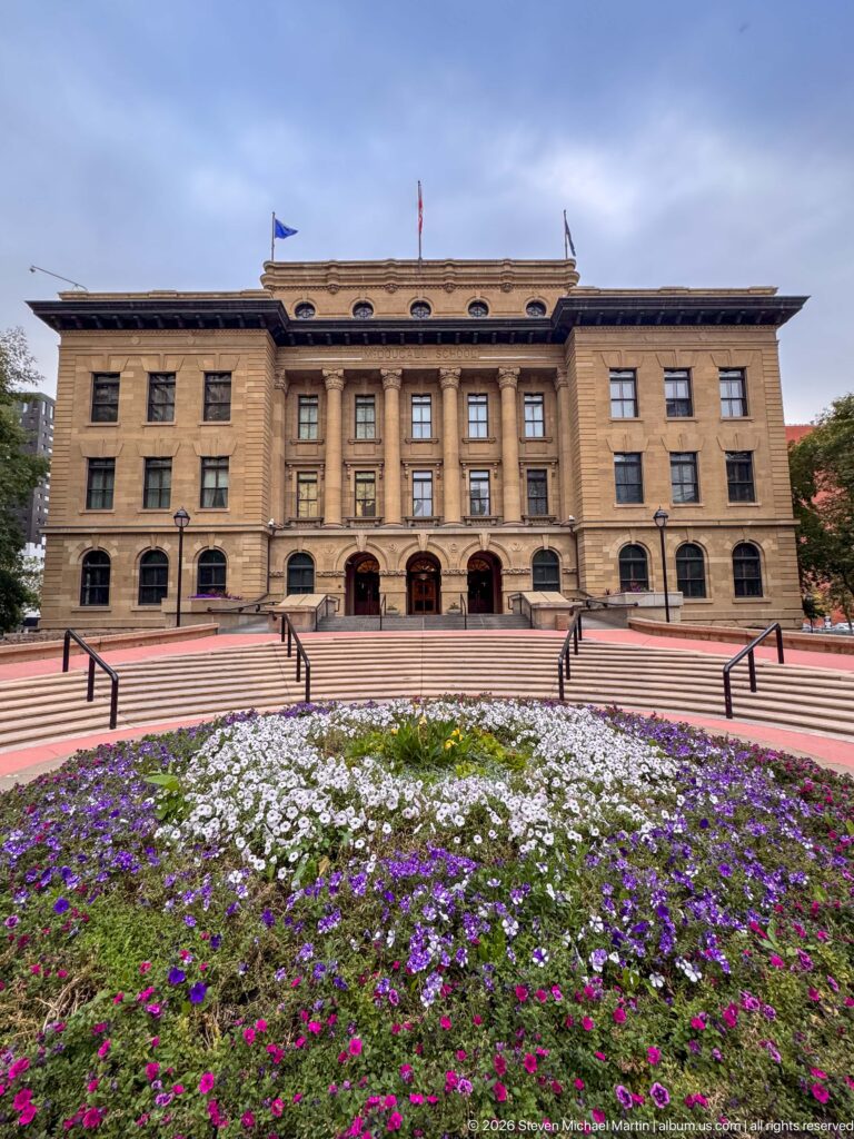 McDougall Centre behind purple, white, and magenta flower beds in Harley Hotchkiss Gardens, with three arched entrances and rooftop flags.