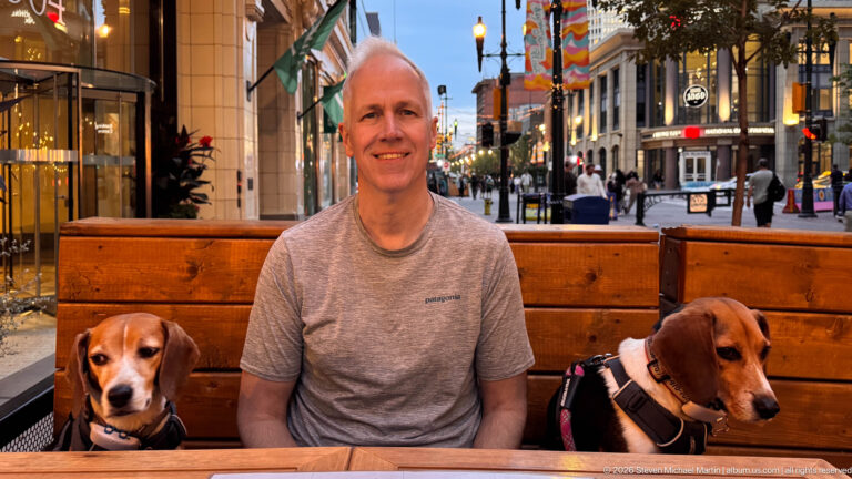 Man seated between two beagles at an outdoor Calgary restaurant patio with illuminated downtown street behind.