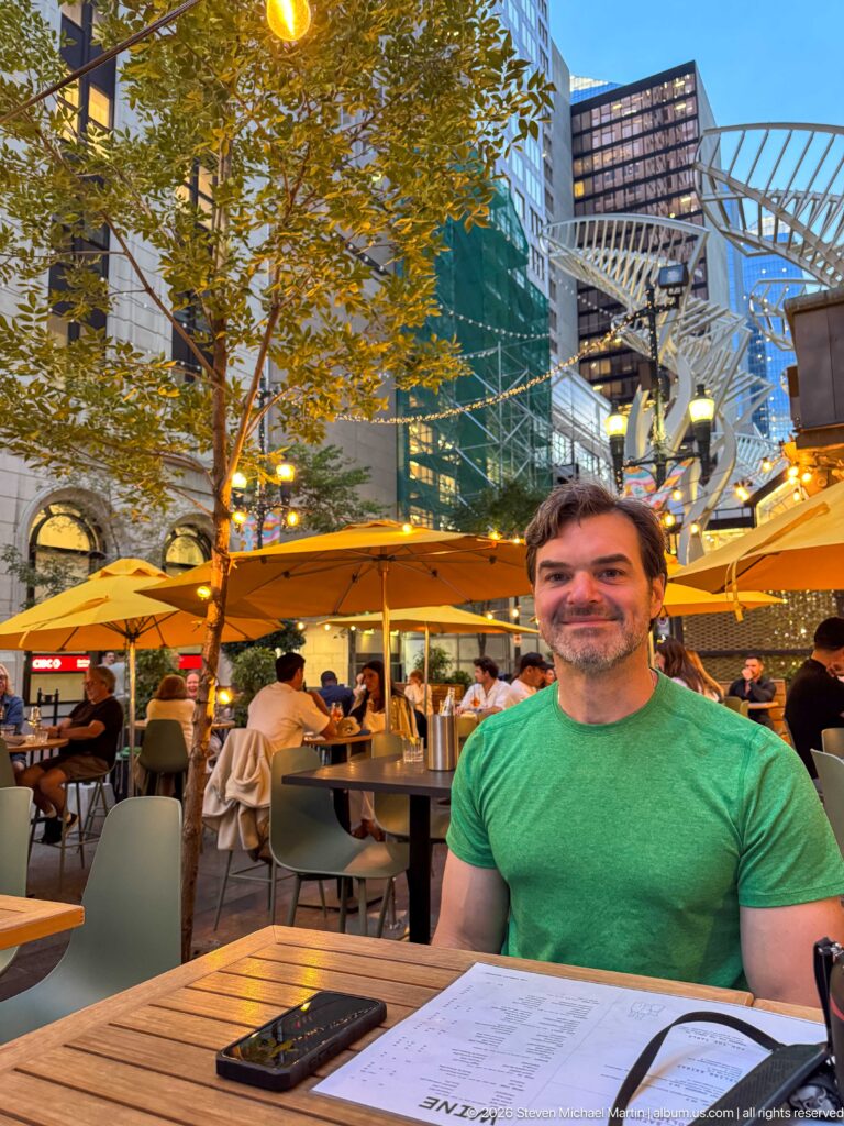 Man in green shirt seated at an outdoor restaurant table beneath yellow umbrellas in downtown Calgary.