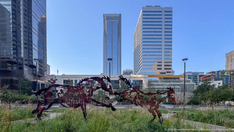 Openwork metal sculptures of running horses among tall grasses with downtown Calgary office towers behind.