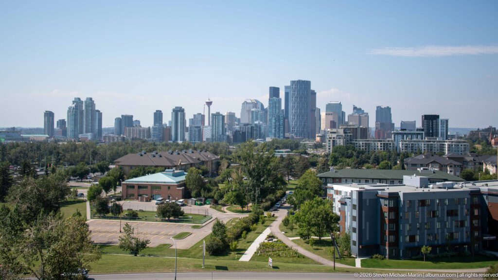 Downtown Calgary skyline beyond green parkland, paths, trees, low buildings, and a modern apartment block.