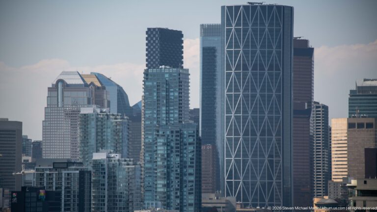 Telephoto Calgary skyline with glass residential towers and a tall dark office building crossed by white diagonal braces.
