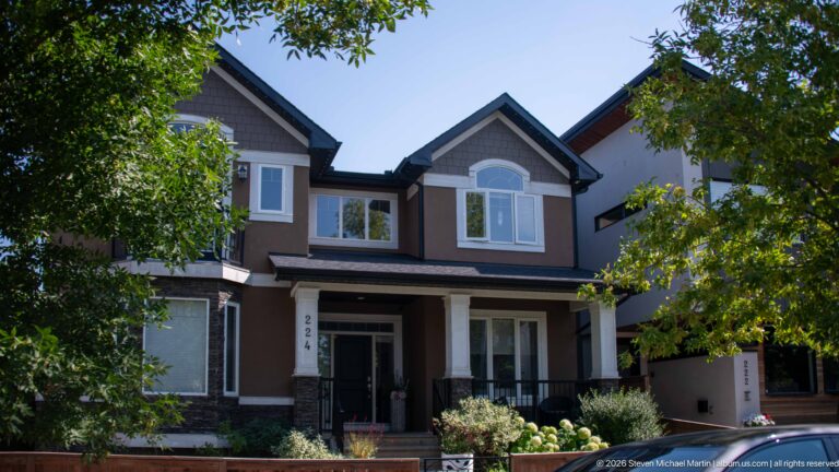 Large contemporary Calgary house with multiple gables, taupe siding, white trim, covered porch, and mature trees.