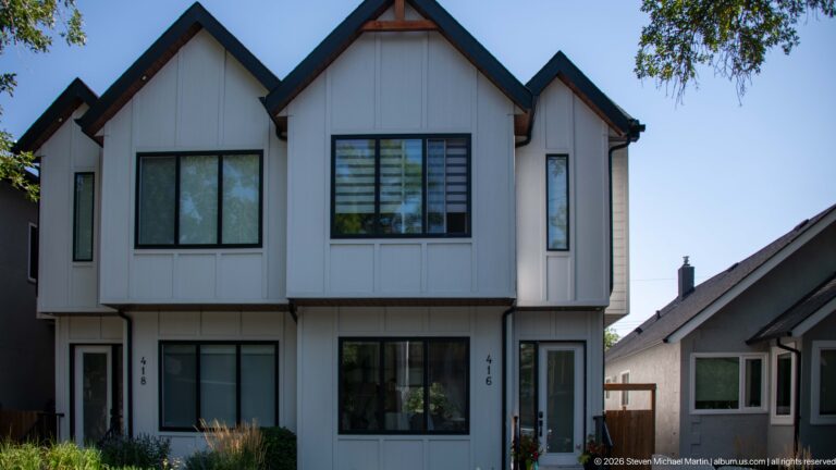 Contemporary Calgary duplex with white vertical siding, black windows, steep gables, wood soffits, and two recessed entrances.