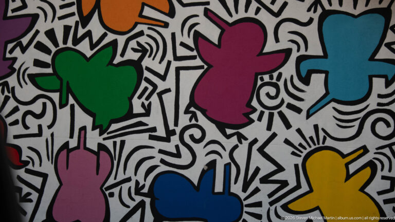Colorful abstract human-like figures outlined in black against a white wall filled with energetic black line patterns.